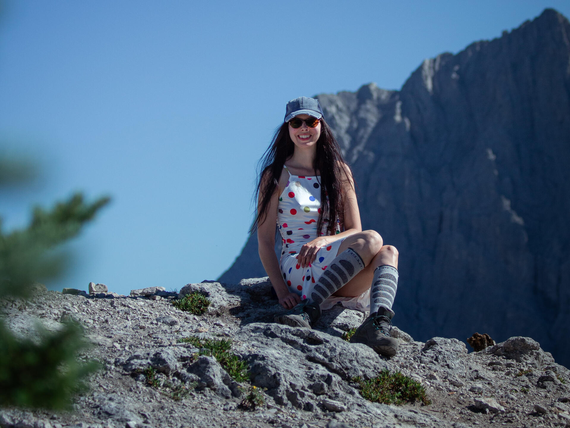 Layla Messner late-diagnosed autistic high-masking autistic creator of the Acute Protocol burnout recovery roadmap A smiling young woman with long brown hair, wearing a polkadot dress, hiking boots, and a ball cap sits on a rock in the alpine